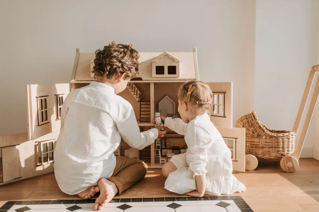 Deux enfants jouent avec une maison de poupée en bois dans une pièce lumineuse